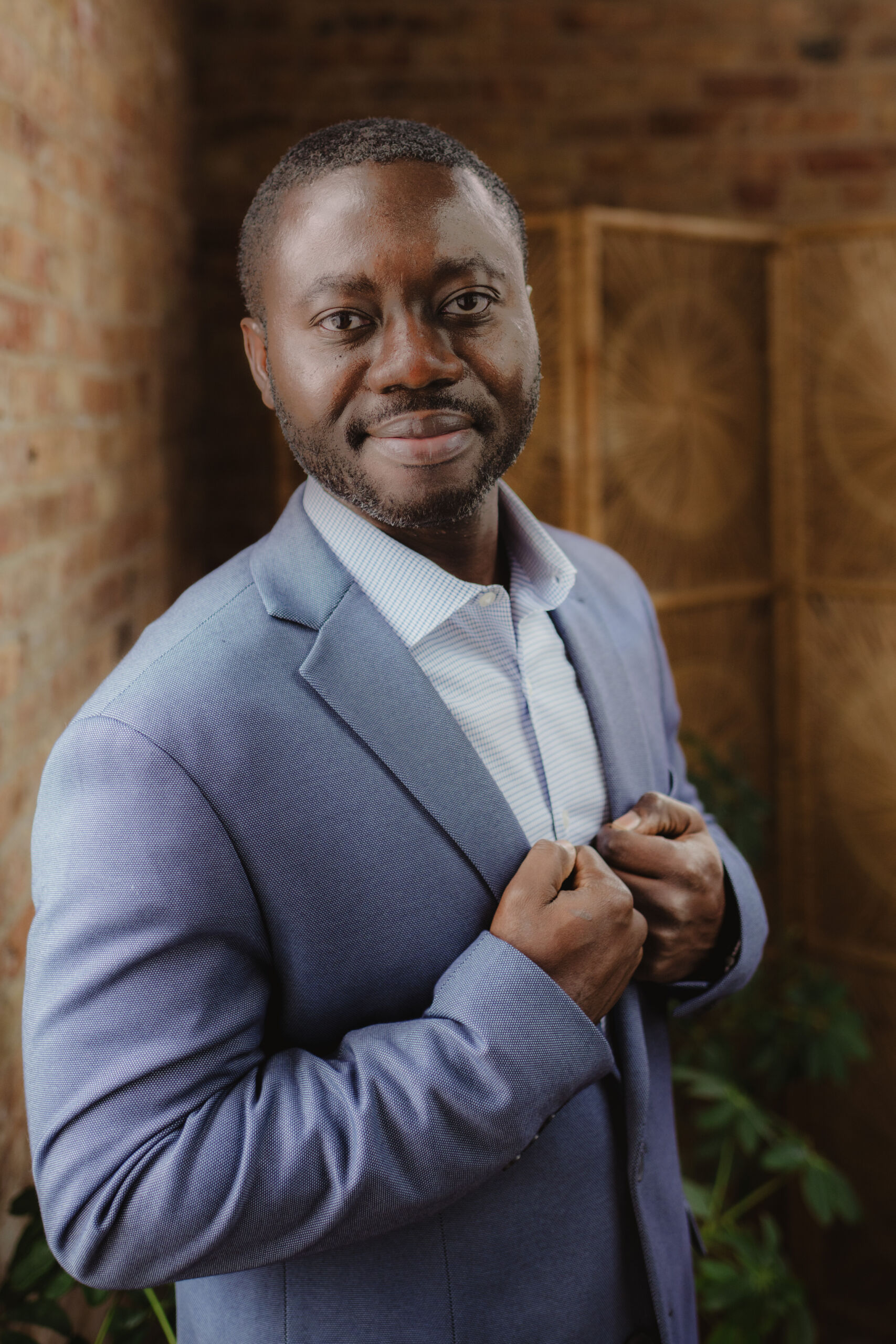 Portrait of a man in a blue suit jacket, standing confidently in a warmly lit room with brick walls and greenery.