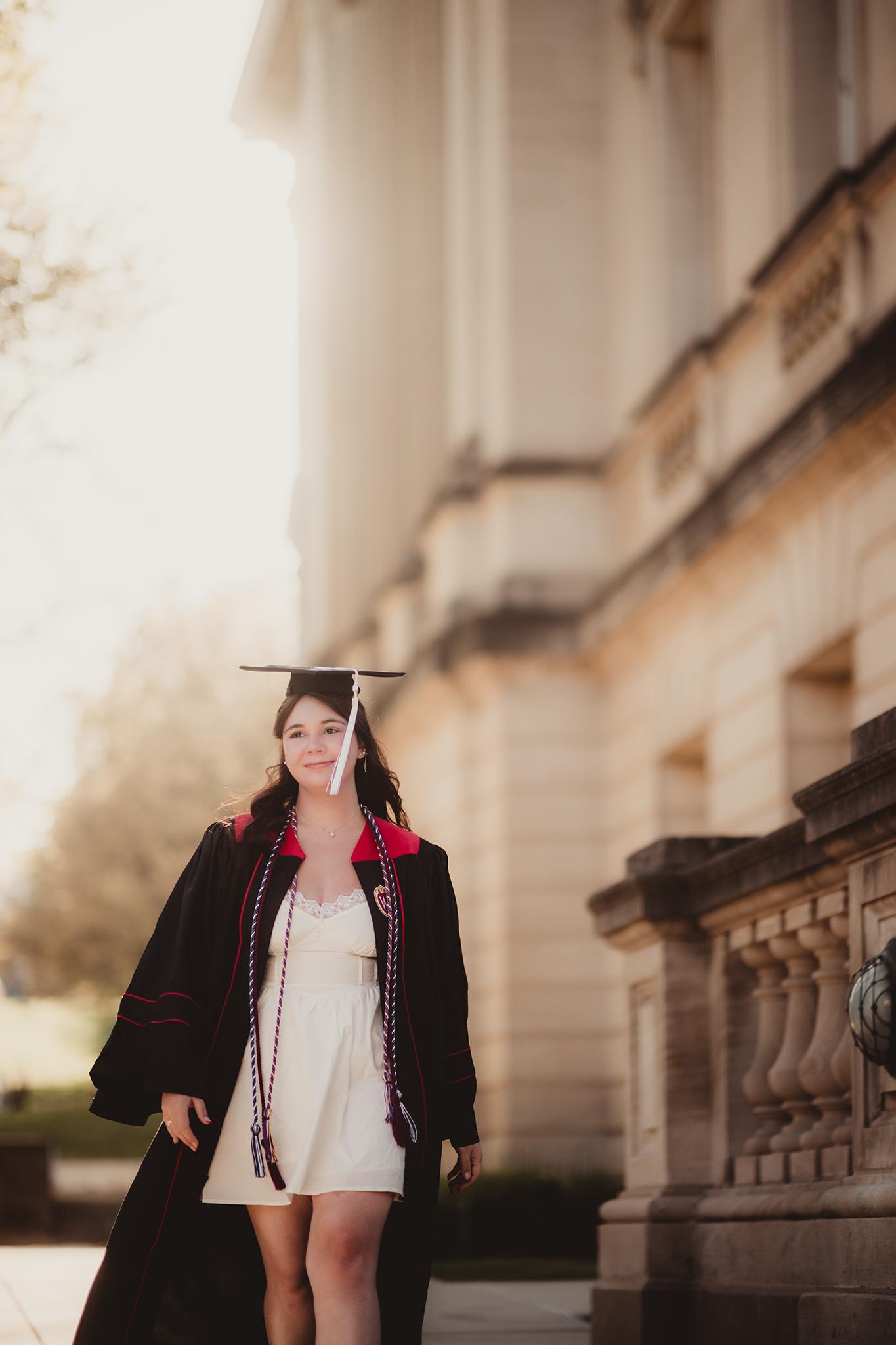 A young woman in graduation attire joyfully walks past a historic building, celebrating her achievement.