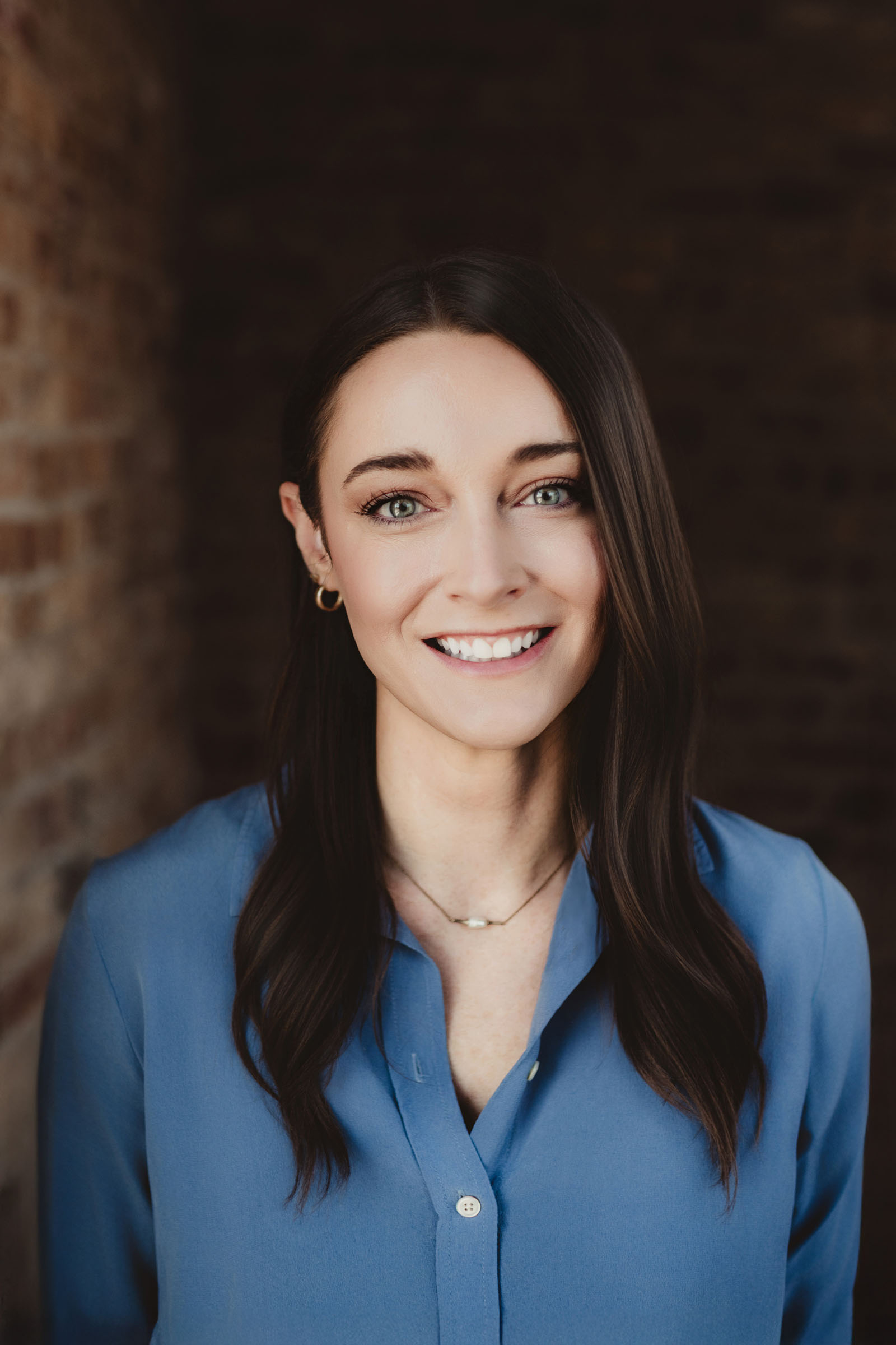 Woman with long dark hair, wearing a blue blouse, smiling warmly against a brick wall backdrop.