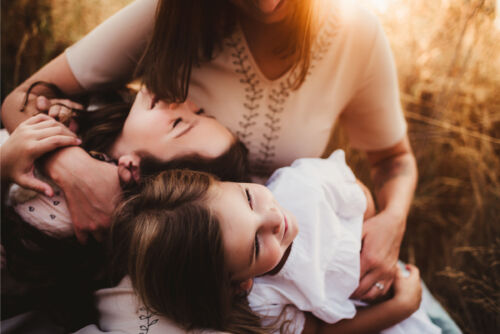 A warm family hug captured in a grassy field at sunset, featuring a mother embracing her two children with genuine smiles.