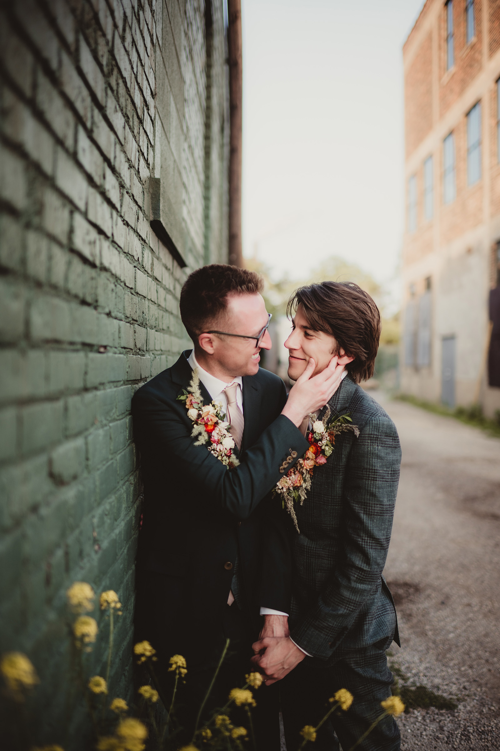 Two grooms sharing a tender moment against a green brick wall with floral accents in their attire.
