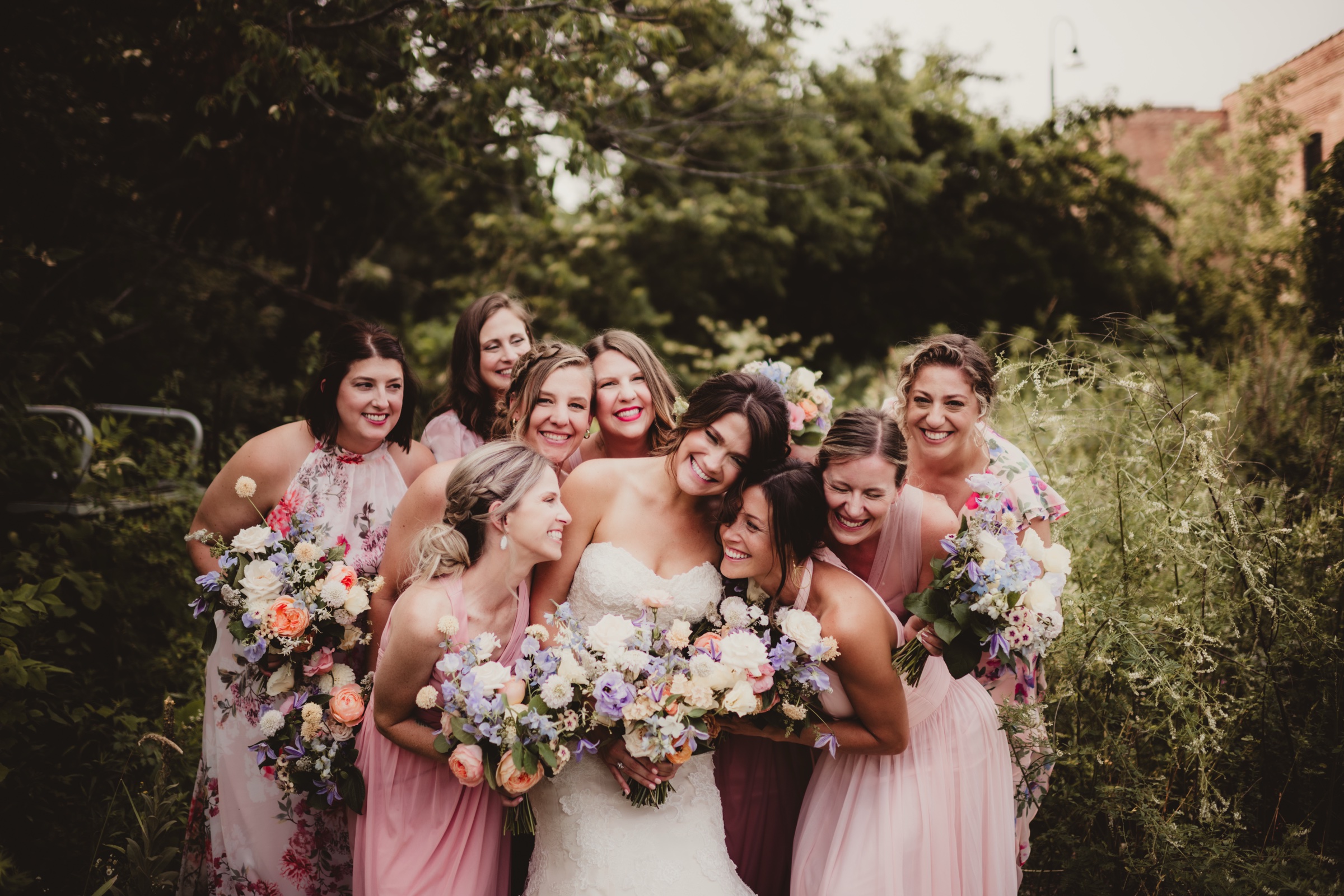 Bride surrounded by bridesmaids with colorful floral bouquets in the outdoor green space at Garver Feed Mill with the building visible behind them