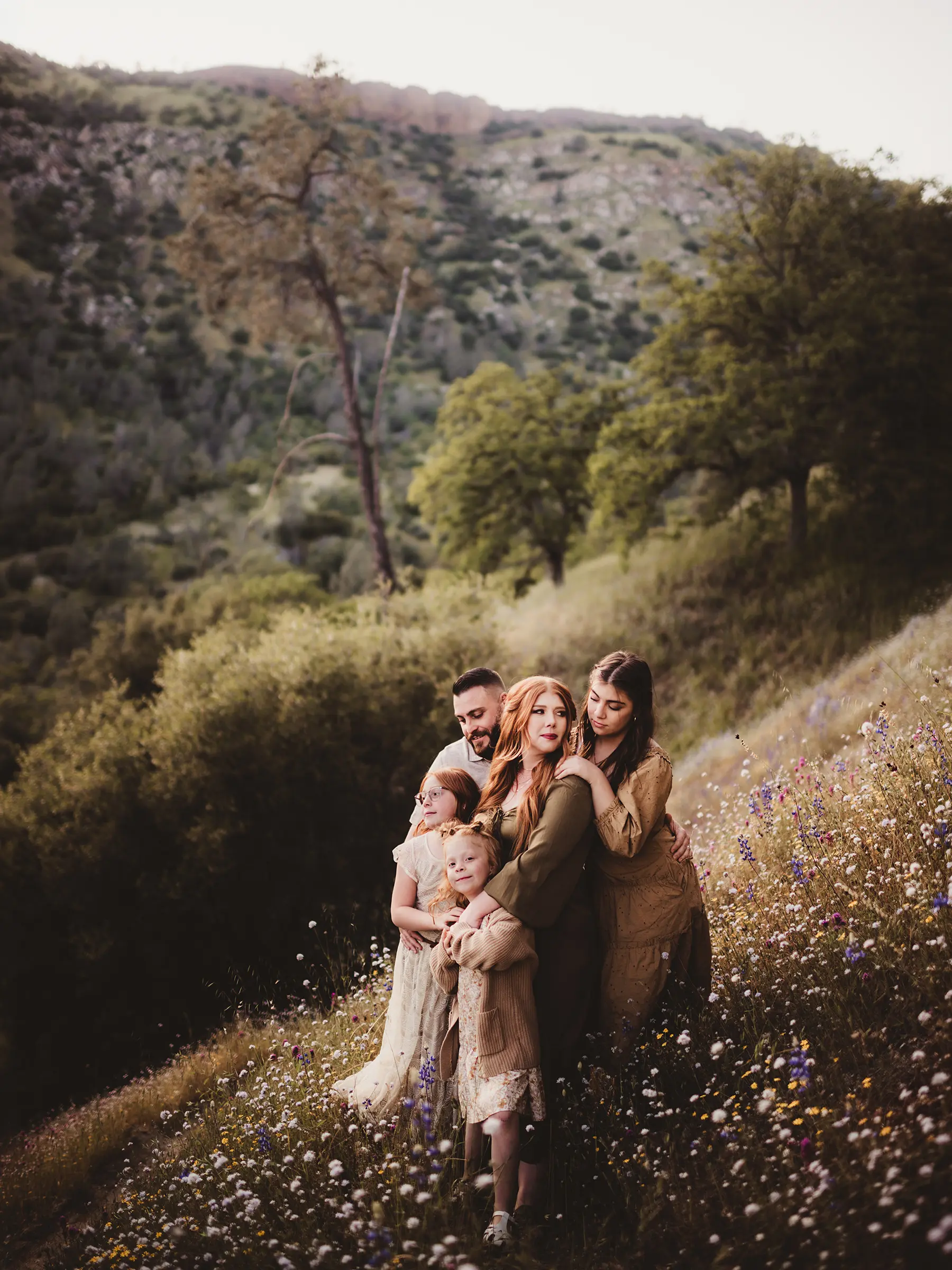 Family of five embracing on a hillside covered with wildflowers surrounded by lush greenery and mountains