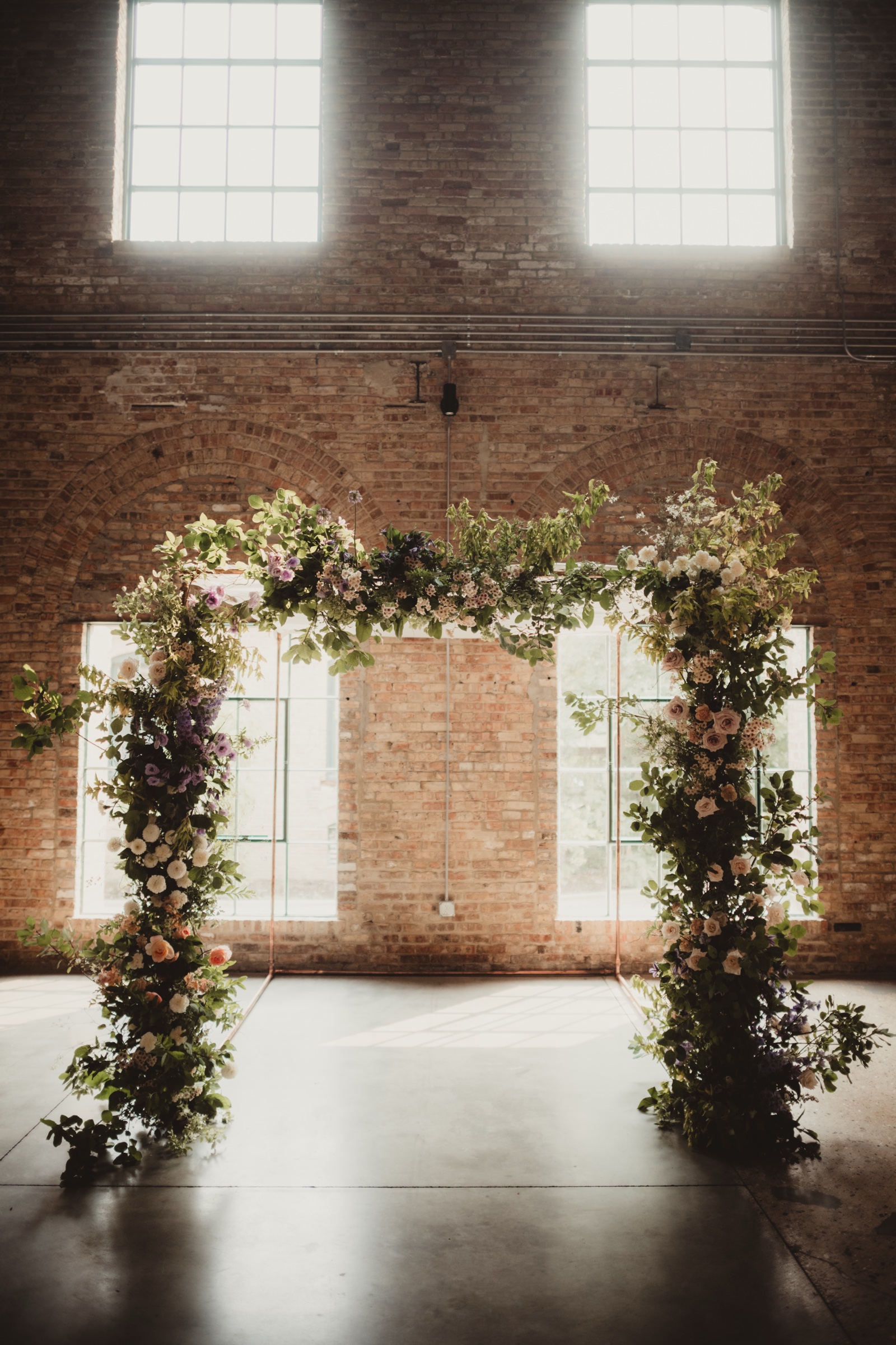Lush floral ceremony arch framed by arched brick windows with soft natural light streaming into the Garver Feed Mill atrium
