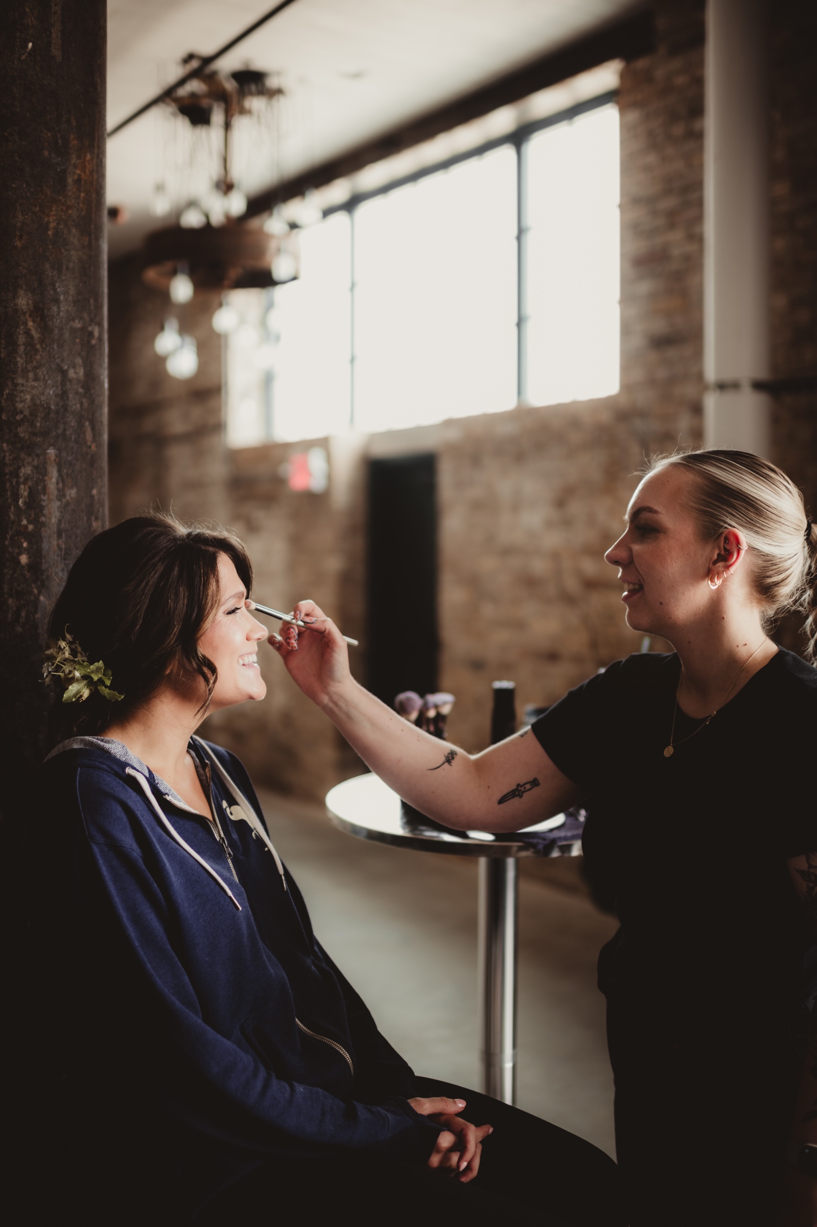 Bride getting makeup done inside the Garver Canvas space with tall industrial windows and exposed brick visible behind the artist