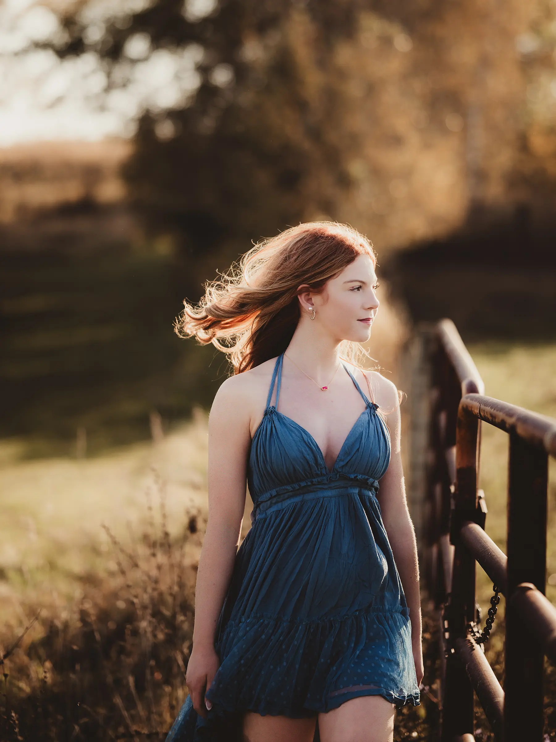 Young woman with red hair in a flowing blue dress walking beside a rustic fence in a sunlit field for senior portrait photography