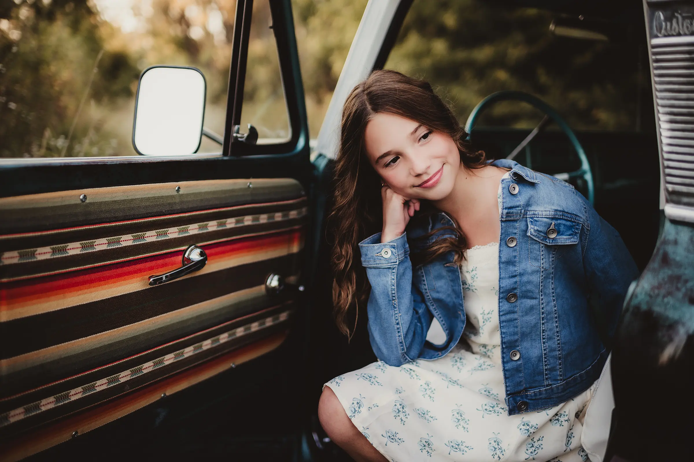 Young girl in a denim jacket and floral dress posing in a vintage car for a portrait session by Twig and Olive Photography
