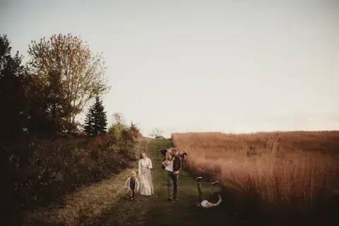 A family plays together along a path between trees and a field of tall grass.