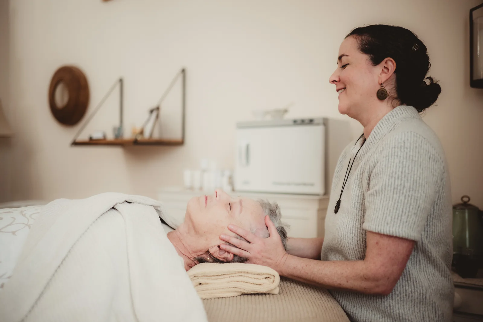 A woman smiles while working beside someone reclining on a treatment table.
