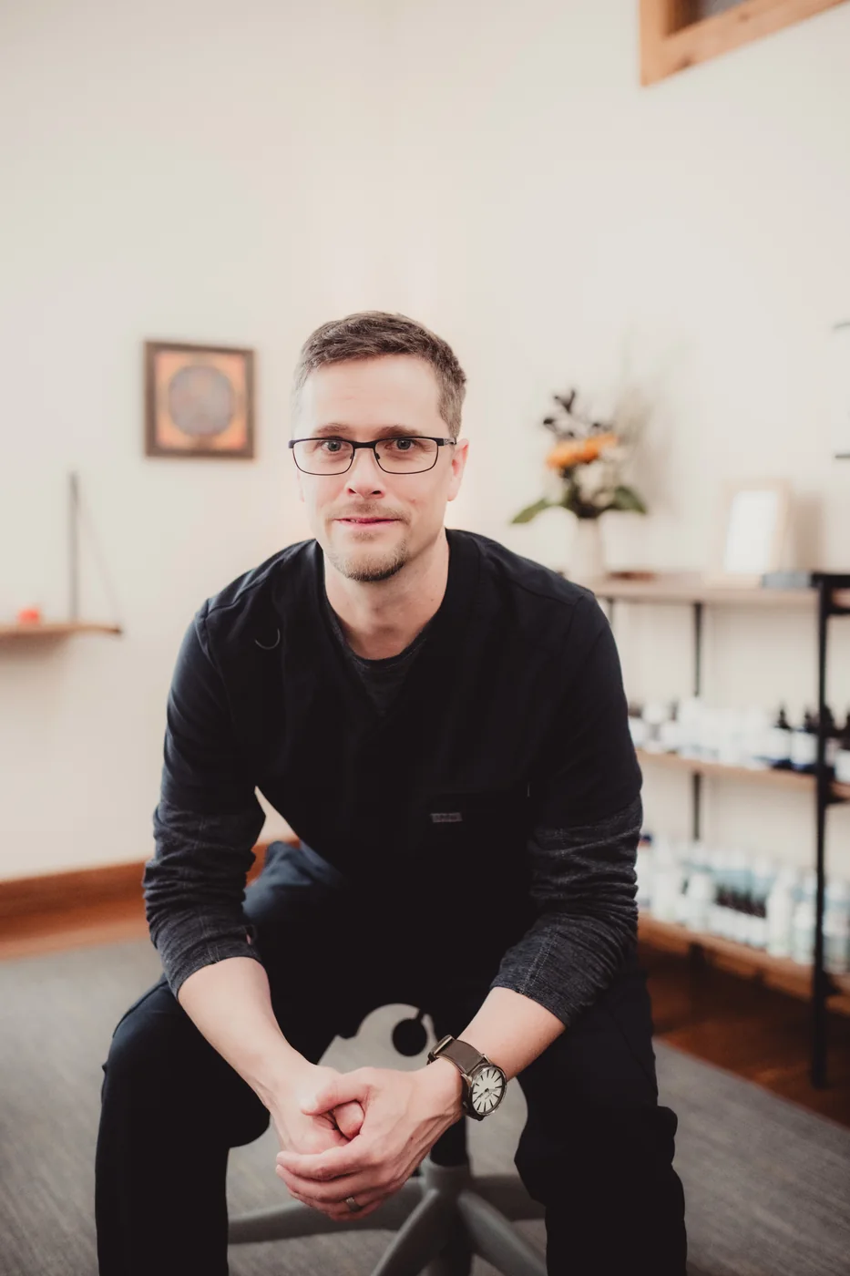 A man wearing glasses sits in a workspace with products arranged behind him.