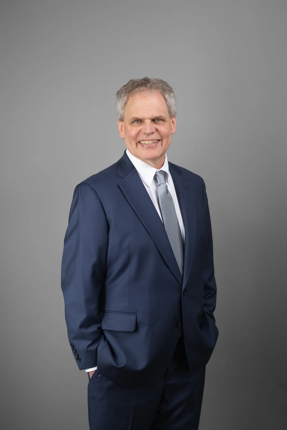 gtw-law-firm-team-headshot-01 Person in a navy suit and pale tie standing against a gray studio background.