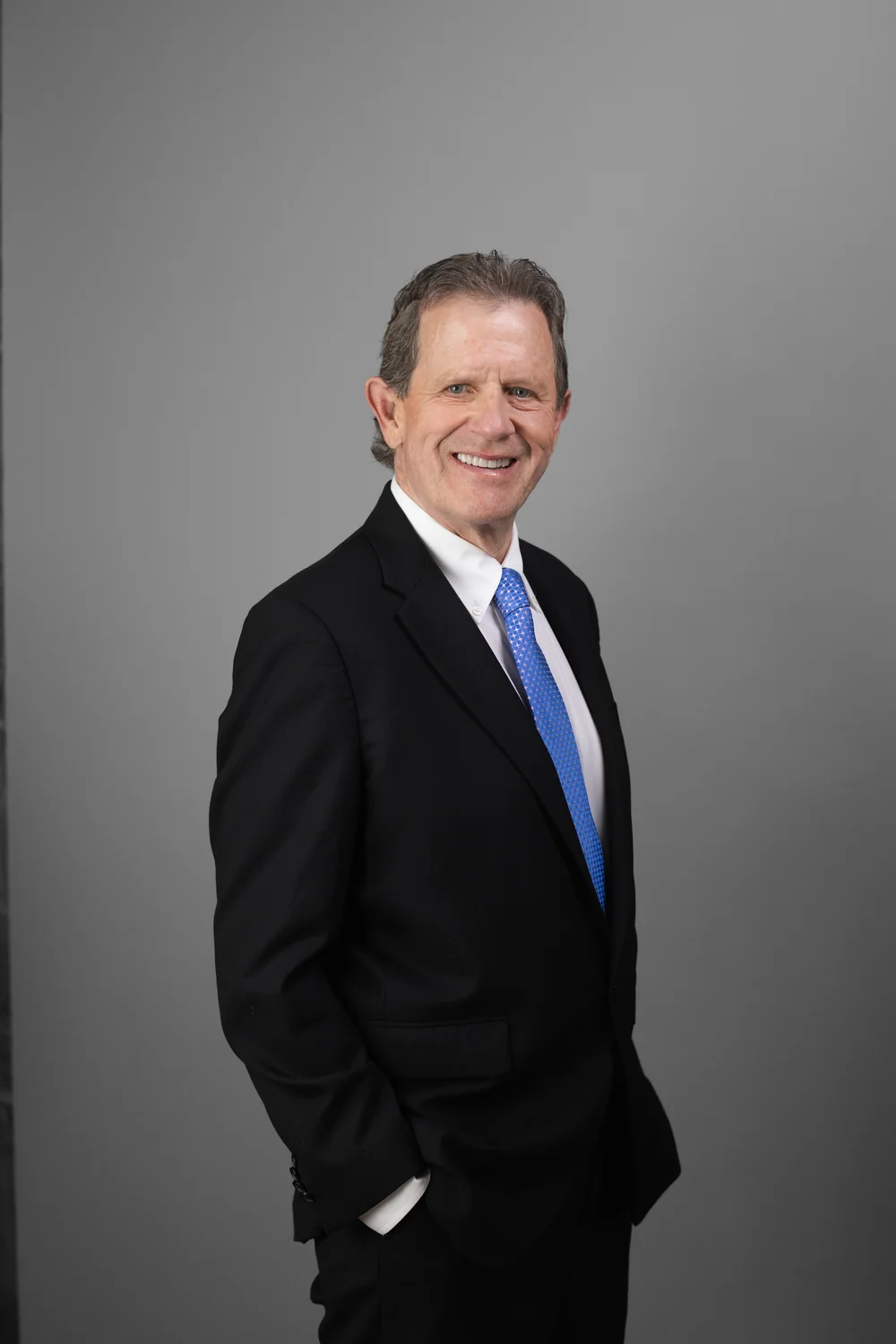 gtw-law-firm-team-headshot-02 Person in a black suit and blue tie with hands in pockets against a gray studio background.