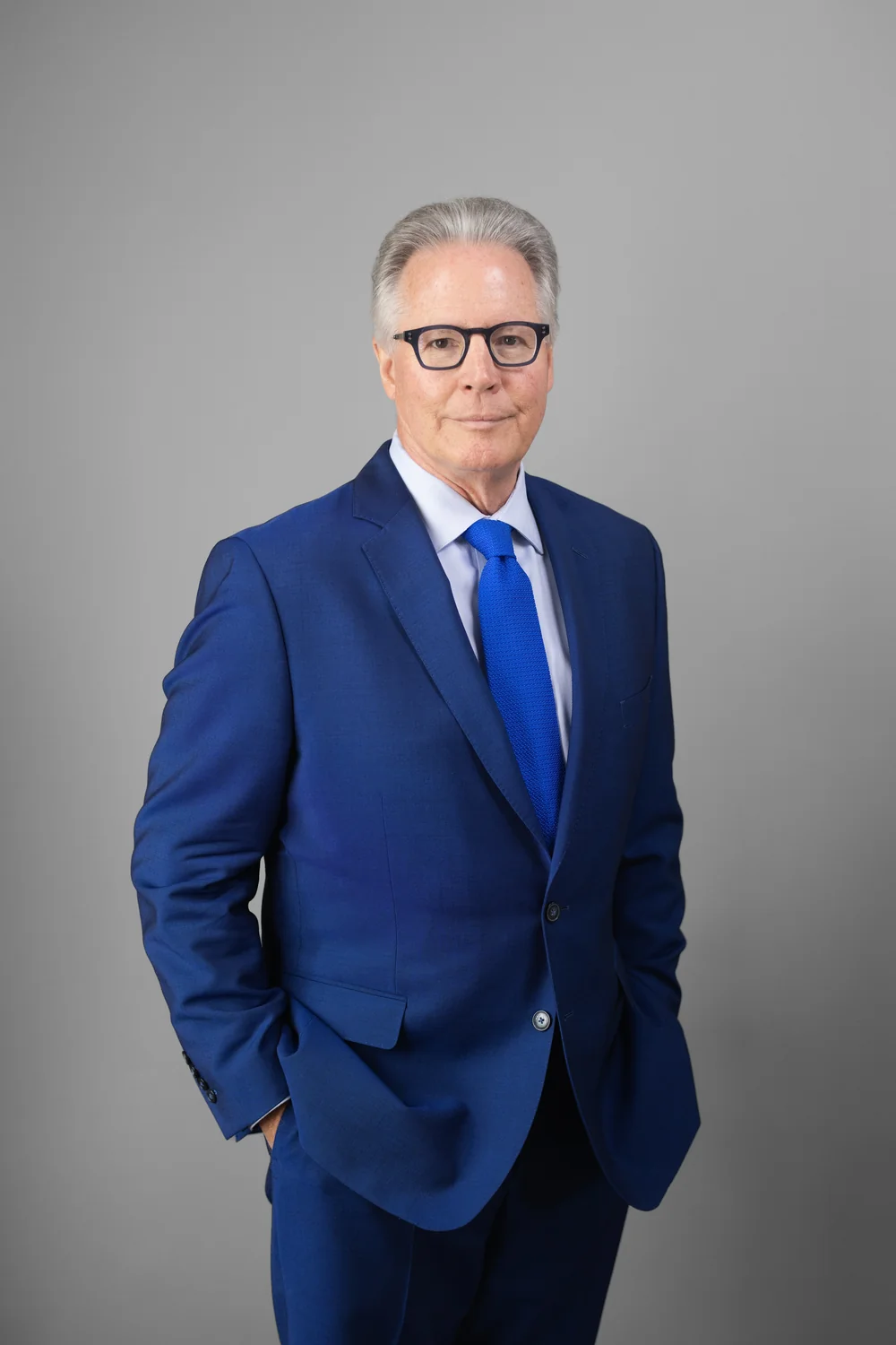 gtw-law-firm-team-headshot-05 Person wearing glasses, a bright blue suit, and a blue tie against a gray studio background.