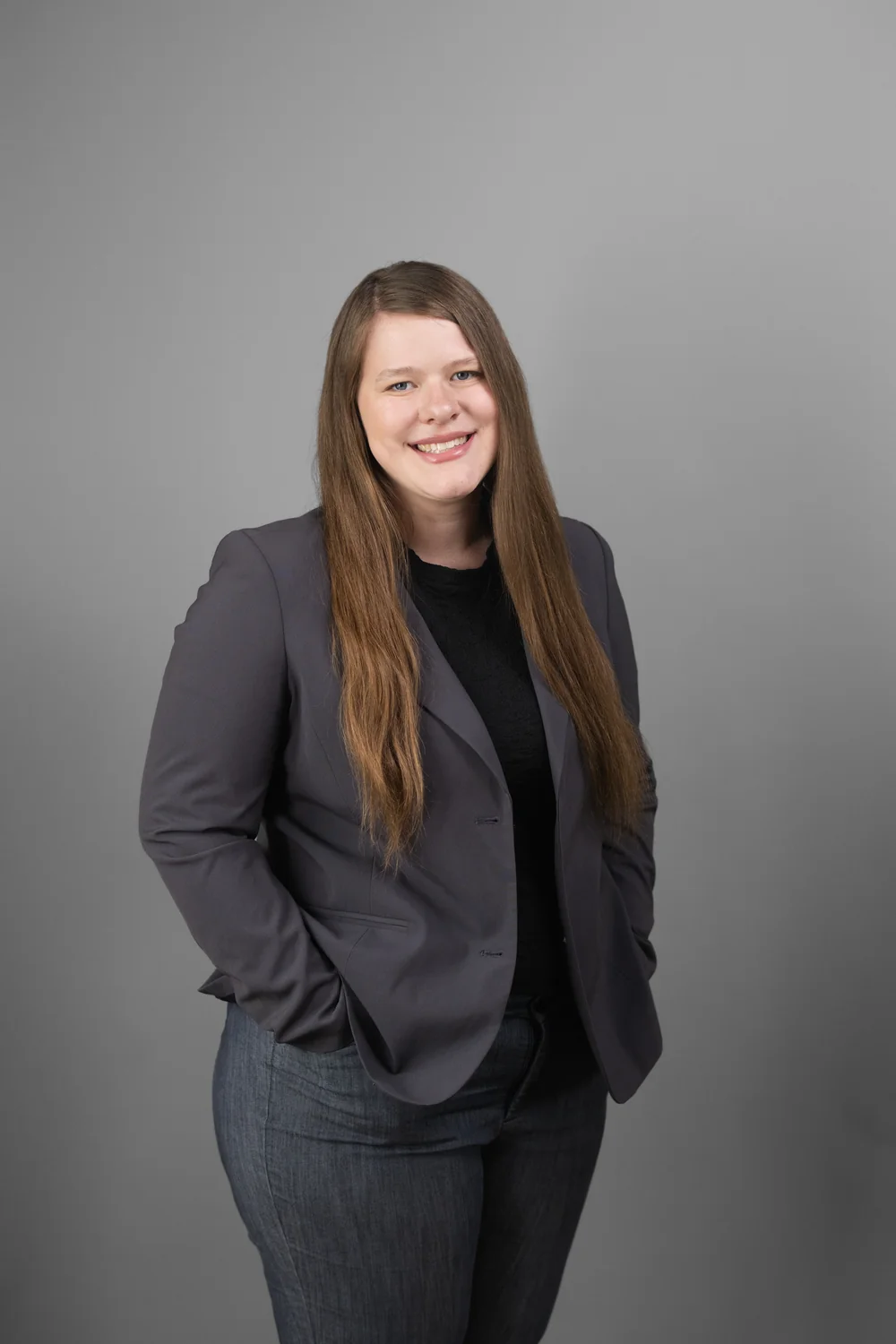 gtw-law-firm-team-headshot-06 Person with long brown hair in a gray blazer and black shirt against a gray studio background.