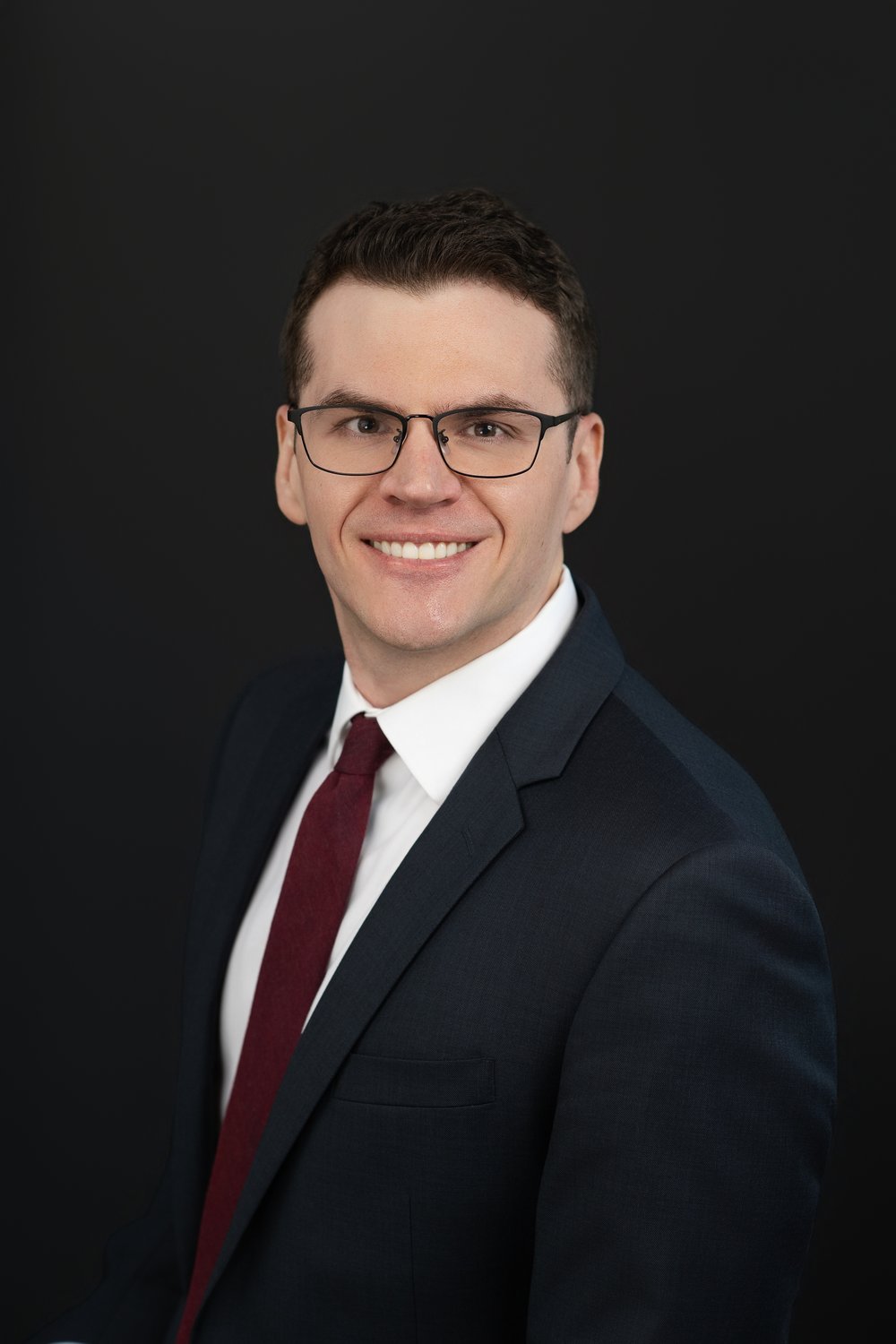 madison-custom-headshot-glenn Smiling man wearing glasses, a navy suit, and a burgundy tie against a dark studio background.