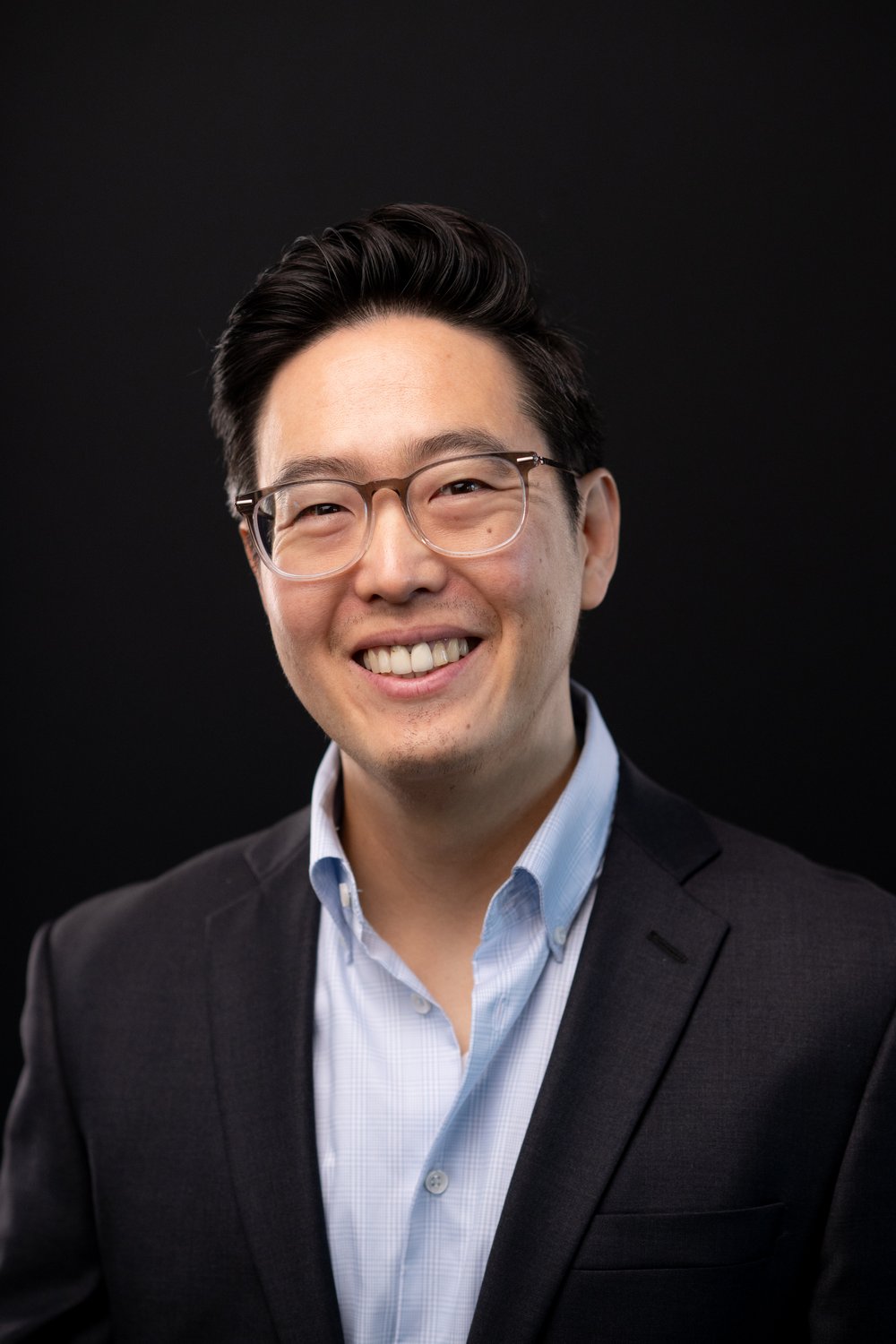 madison-custom-headshot-mike Smiling man wearing glasses, a dark blazer, and a light blue shirt against a dark background.