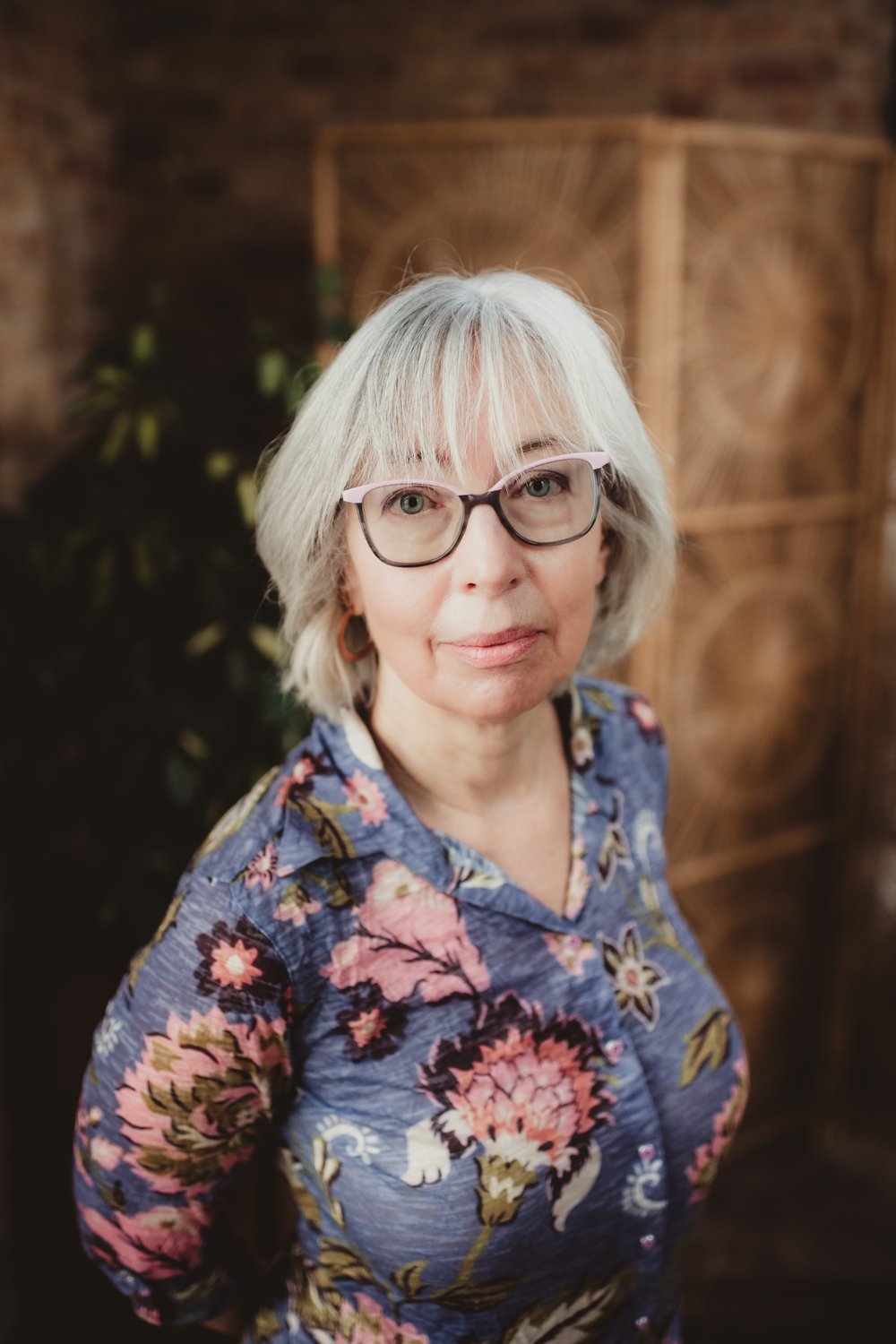 madison-drop-in-headshot-mona Woman wearing glasses and a floral blouse against a softly blurred brick and wood background.