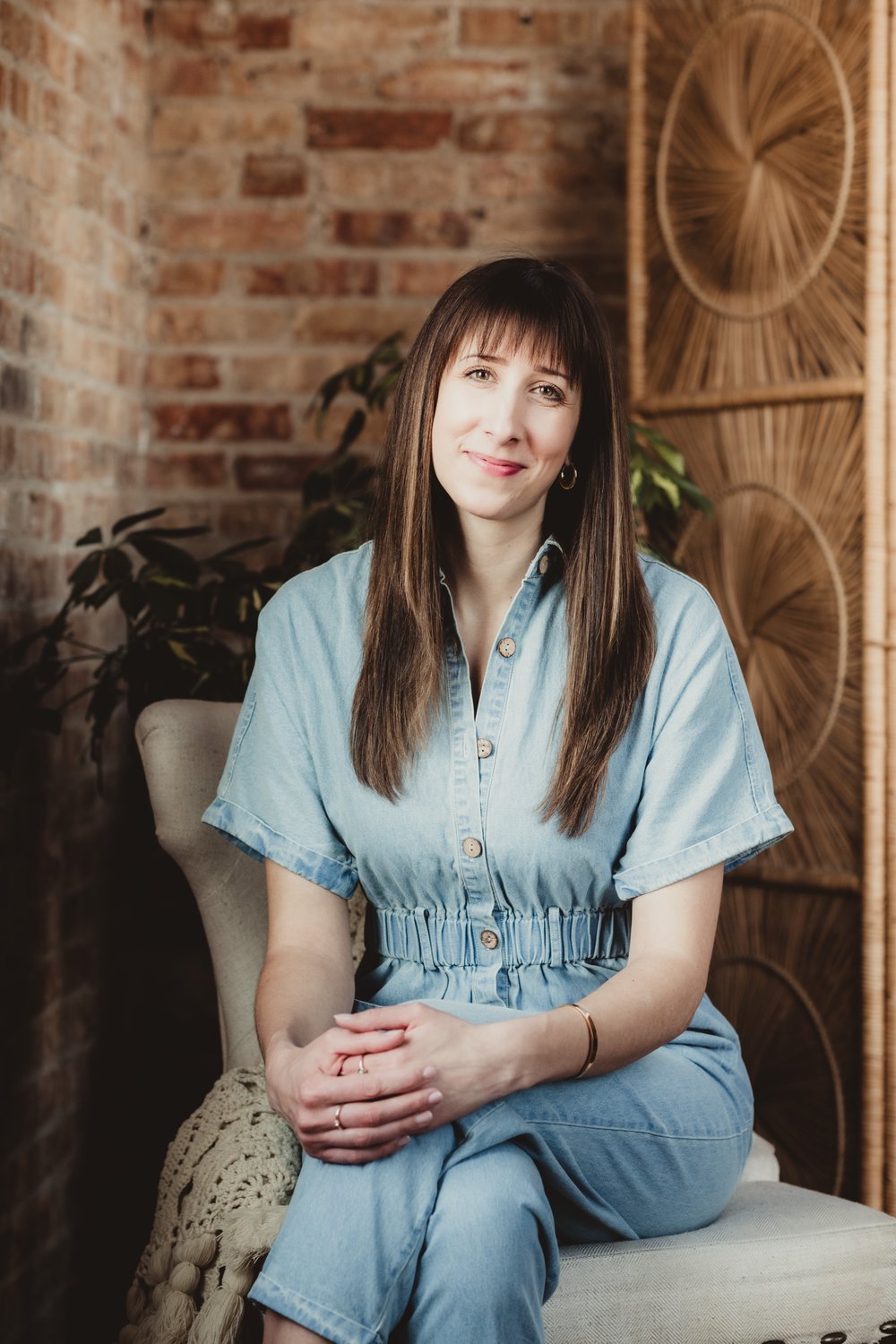 madison-drop-in-headshot-samantha Woman in a light blue outfit seated in a studio with exposed brick, plants, and a woven screen.