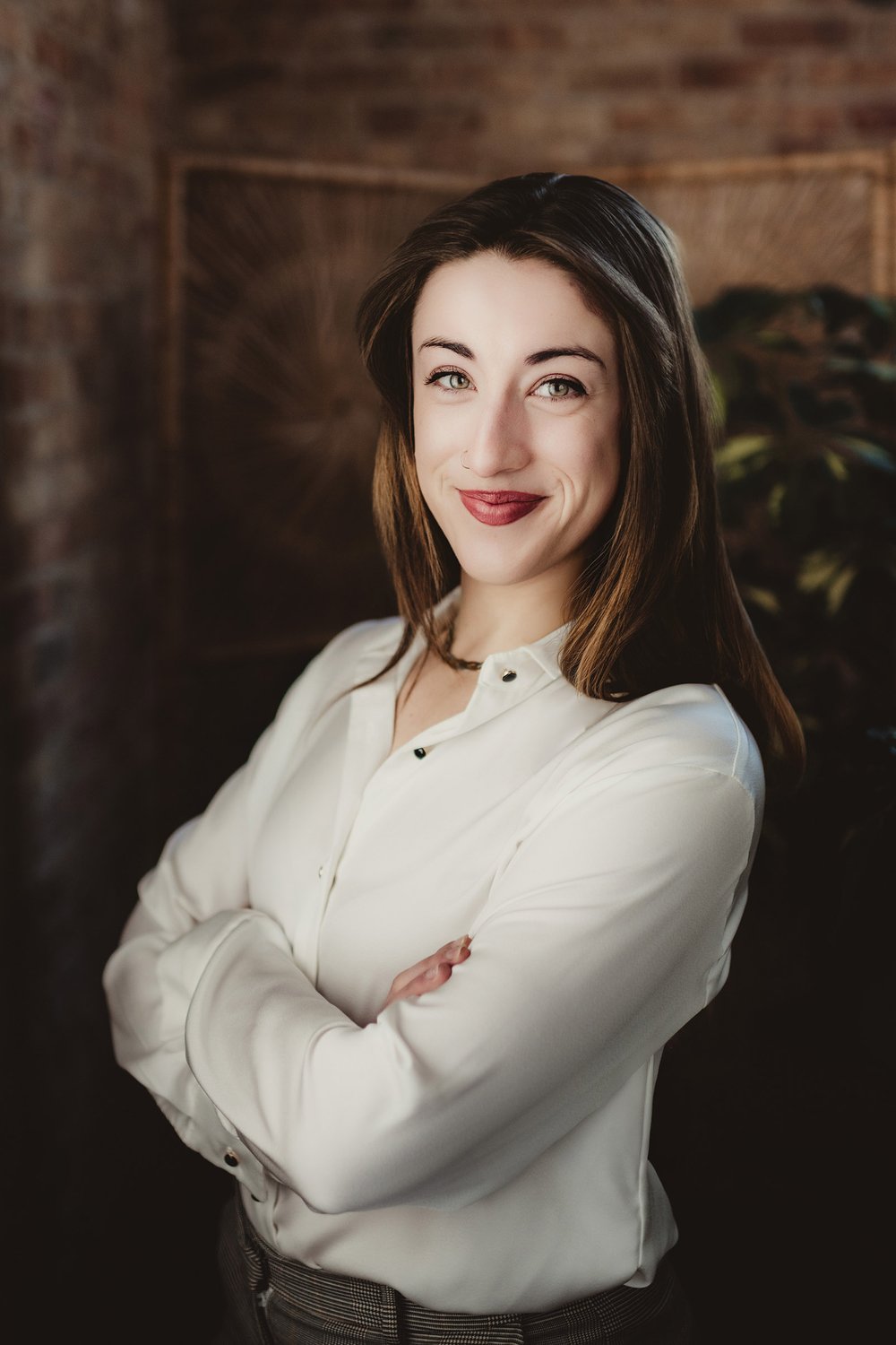 madison-drop-in-headshot-zoe Woman in a white blouse with her arms crossed beside exposed brick and a wooden door.