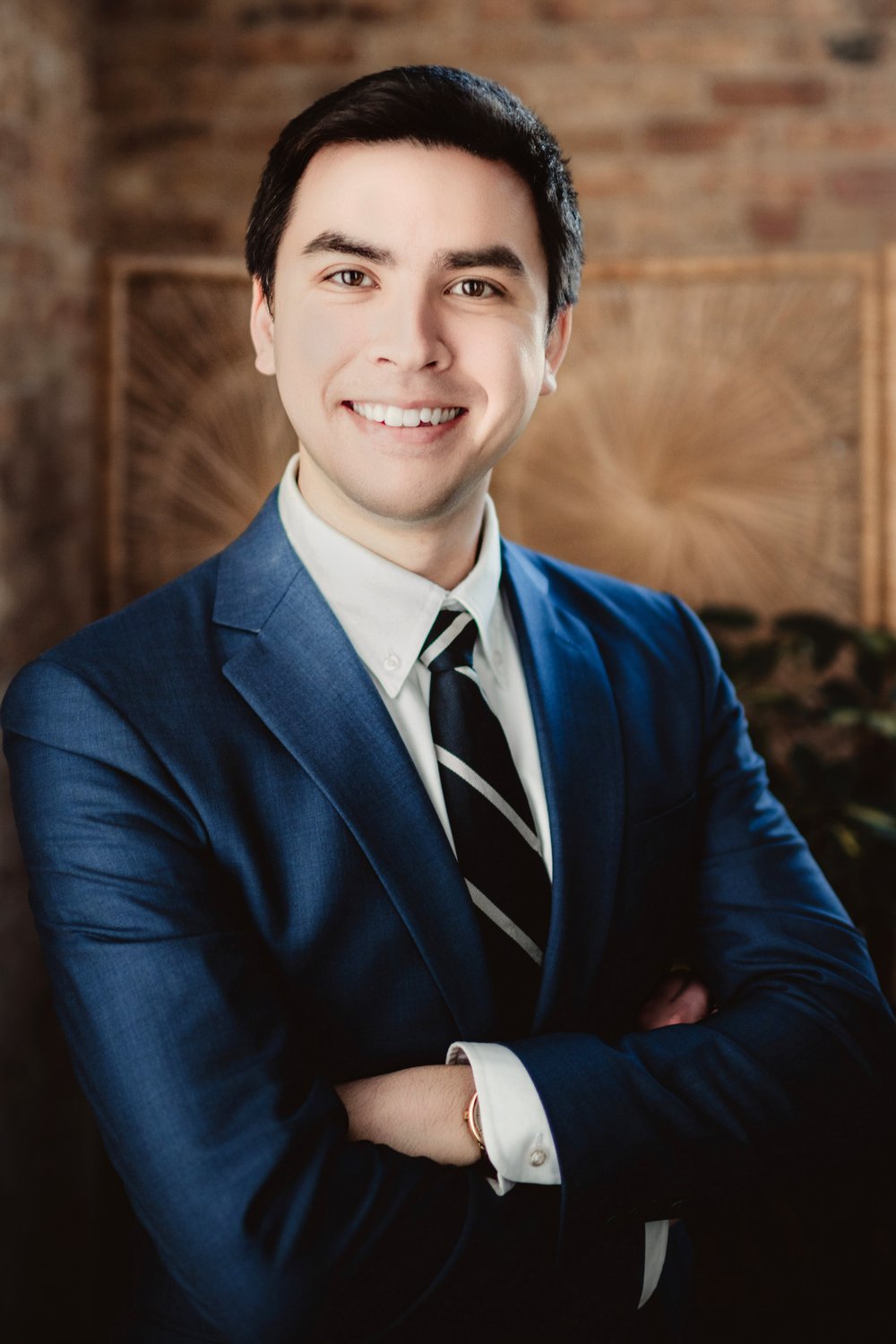 madison-studio-headshot-niall Smiling man in a blue suit and striped tie with his arms crossed in front of brick and wood.