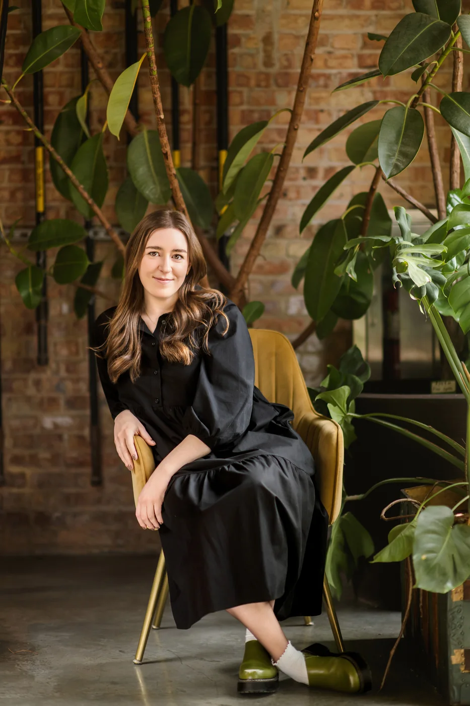 A woman in black sits on a chair among tall plants beside an exposed brick wall.
