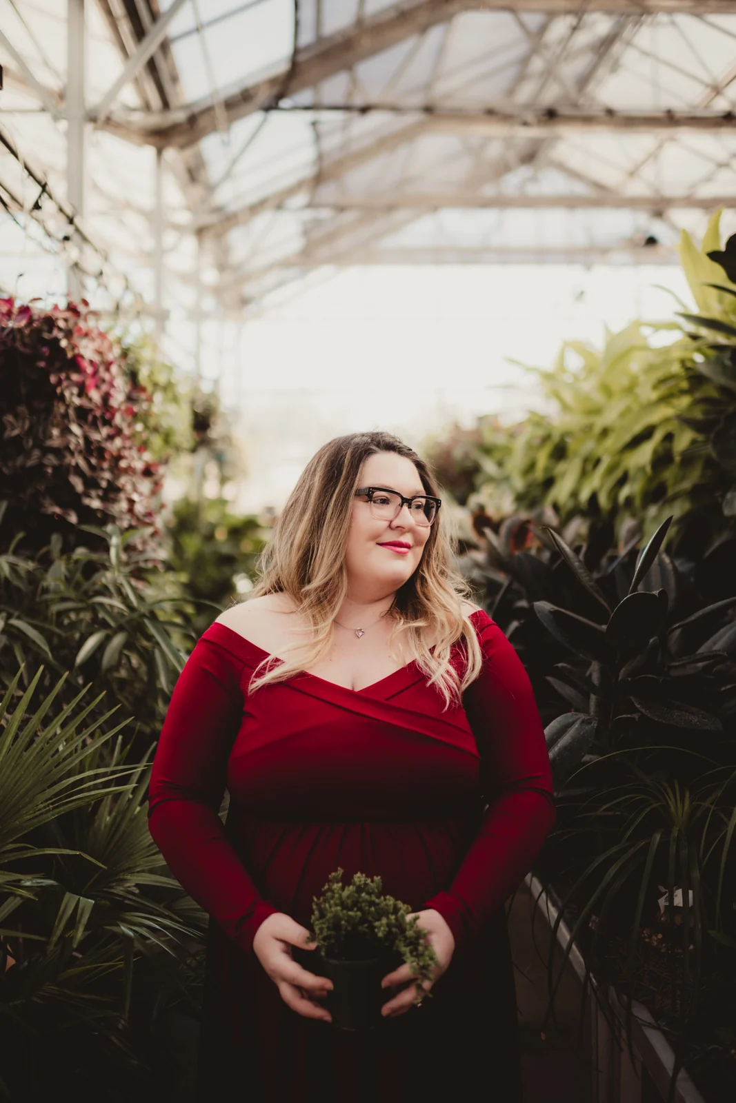 Maternity portrait in a greenhouse A pregnant woman in a red dress standing among greenhouse plants.