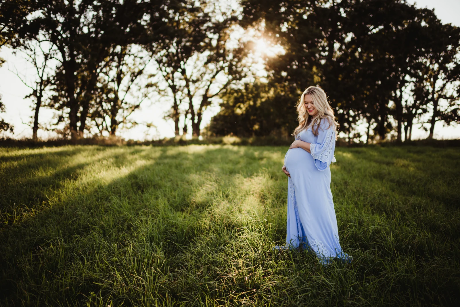 Maternity portrait on a sunlit hillside A pregnant woman in a pale blue dress standing on a sunlit grassy hillside.