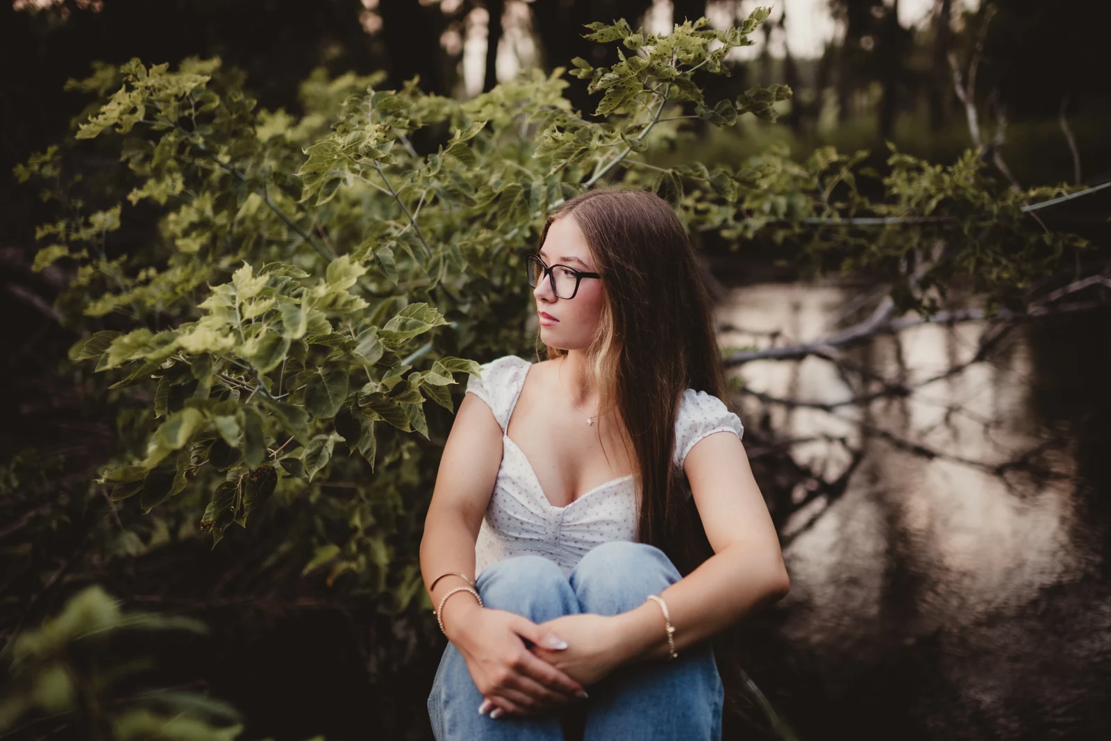 to-senior-2026-senior-woodland-portrait A senior wearing glasses seated among green woodland foliage.