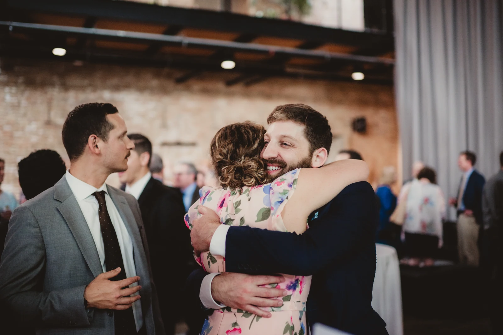 Person in a navy suit hugging a guest in a floral dress at the reception.